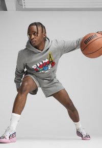 Grijze hoodie met kleurrijk grafisch logo, bijpassende grijze shorts en roze sneakers met cartoonpatronen. Houdt een basketbal vast.