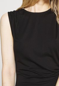 Top sans manches noir avec détails froncés aux épaules, en tissu lisse, présentant une silhouette ajustée et un col rond.