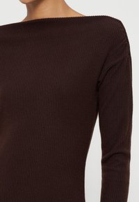 Femme portant un pull en tricot côtelé marron foncé avec des manches longues et un large col bateau.