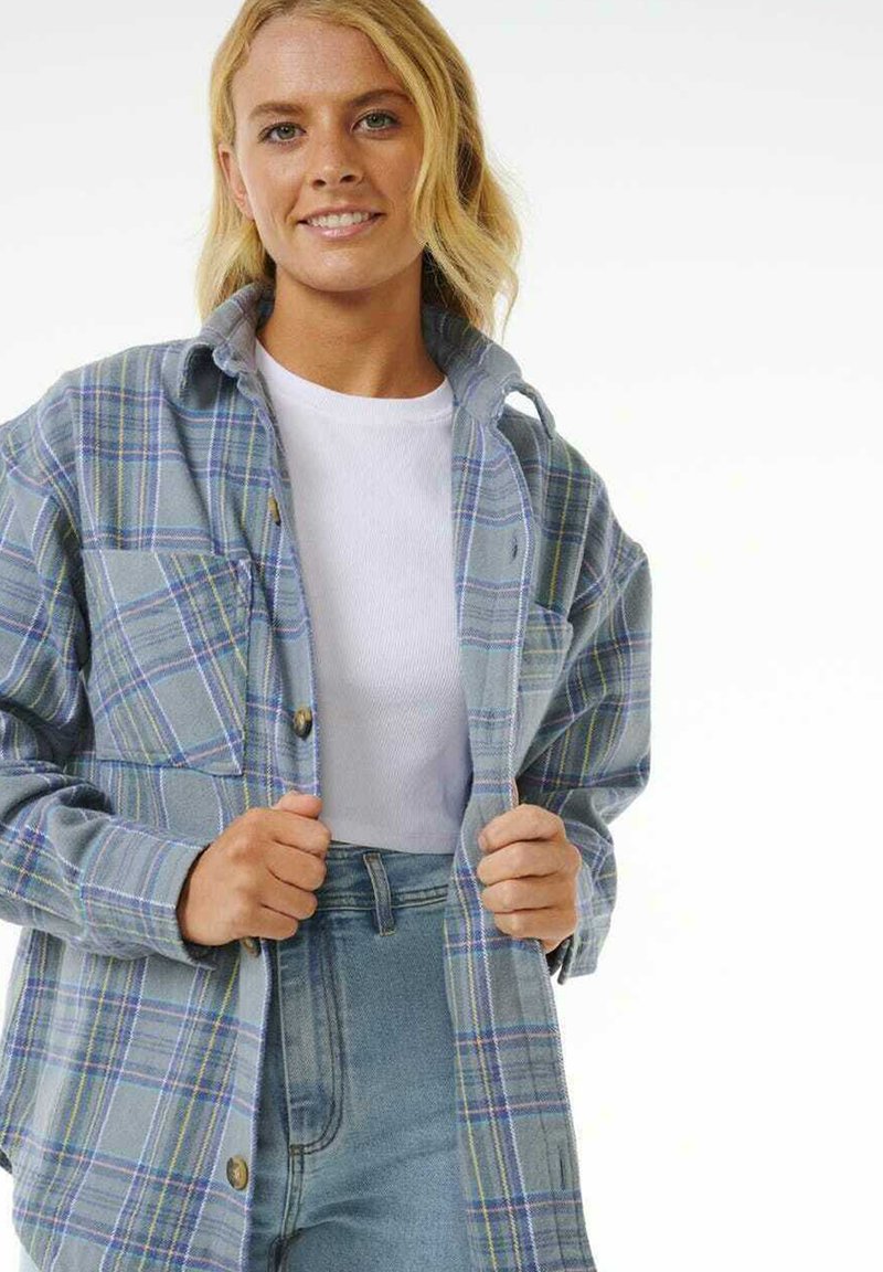 Camisa de franela de cuadros azul y gris con dos bolsillos en el pecho y cierres de botón, usada sobre una camiseta blanca y jeans de cintura alta de color azul claro.