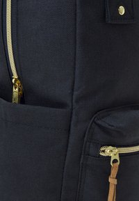 Svart tygryggsäck med guldiga detaljer, en texturerad yta och flera dragkedjefickor med läderaccenter.