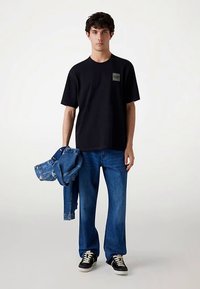 Zwart T-shirt met korte mouwen en een logopatch, gecombineerd met blauwe spijkerbroek. Het model houdt een spijkerjasje vast en draagt zwarte sneakers.