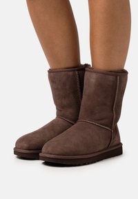 Bruine suède enkelboots met een schapenvachtvoering, gestikte details en platte rubberen zolen, met een eenvoudig, rond teenontwerp.