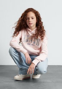 Šviesiai rožinė su kapuci megztinis su užrašu "Levi's EST. 1873" gėlėmis dekoruotu šriftu. Dėvima su šviesiai mėlynomis džinsomis ir baltais batais su raudonu akcentu.