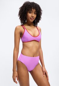 Γυναίκα με σγουρά μαλλιά που φοράει ροζ μπικίνι με κόκκινα περιθώρια, στέκεται μπροστά σε ένα απλό ανοιχτό φόντο, στραμμένη ελαφρώς προς τα δεξιά.