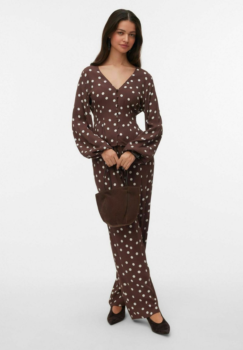 Femme portant une blouse à pois marron et un pantalon, tenant un sac à main marron, debout jambes croisées sur fond blanc.