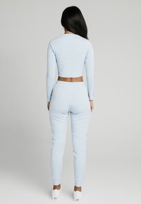 Nainen, jolla on pitkät tummat hiukset, pukeutuneena vaaleansiniseen lyhytkäyttöiseen pitkähihaiseen puseroon ja siihen sopiviin verkkahousuihin valkoisilla lenkkareilla, selkä kameraan päin.