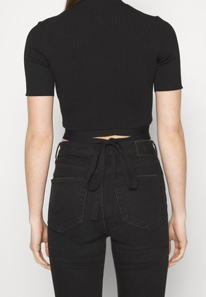 Osoba ubrana w dopasowany czarny prążkowany krótki crop top związany w talii, w połączeniu z czarnymi, wysokim stanem jeansami od tyłu.