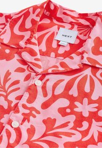 Gros plan d'une chemise à col avec des motifs floraux abstraits rouges et roses et une patte de boutonnage blanche à l'avant, montrant une étiquette de la marque Next.