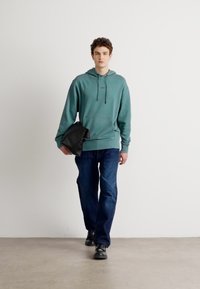 Felpa con cappuccio verde menta e tasca frontale, abbinata a jeans blu scuro. Il modello tiene una borsa nera e indossa scarpe nere. Sfondo semplice.