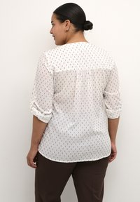 Blusa blanca con pequeños patrones negros, con mangas tres cuartos arremangadas y un sutil detalle de pliegue en la parte posterior.