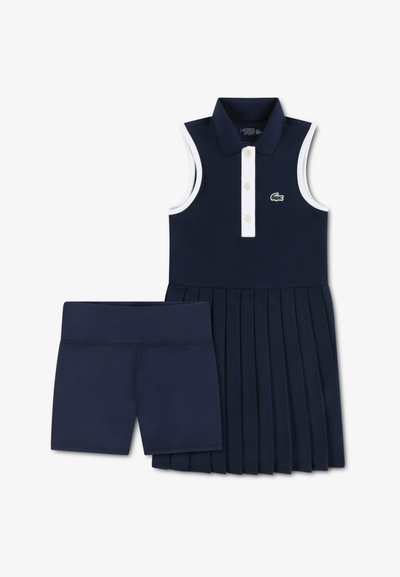 Μπλε σκούρο αμάνικο φόρεμα πόλο με λευκές λεπτομέρειες και πλισέ φούστα, συνδυασμένο με ταιριαστά μπλε σκούρα σορτς, με μικρή πράσινη λεπτομέρεια λογότυπου.