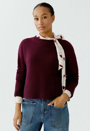 Bordeaux gebreid sweater met een geribbelde zoom en manchetten, met een voorzak. Gecombineerd met een witte sjaal met rode accenten en blauwe jeans.