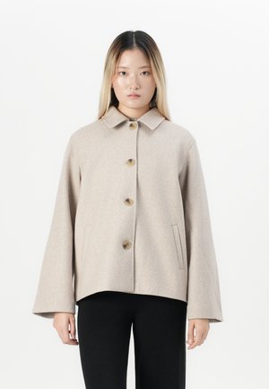 Jeune femme avec des cheveux raides et longs portant une veste beige à boutons avec des manches larges et un pantalon noir sur un fond blanc.