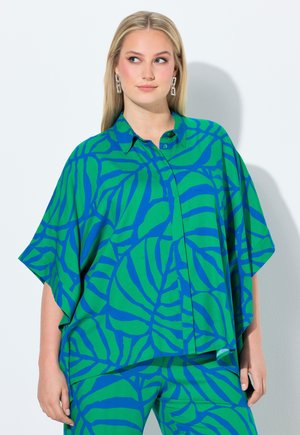 Kvinde iført en løs skjorte og bukser med grønt og blåt bladmønster, poserer med hovedet let vendt til siden.