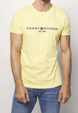 Homme portant un T-shirt ajusté de couleur jaune pâle Tommy Hilfiger avec le logo et le texte "EST. 1985", associé à un jean bleu, sur un fond uni.