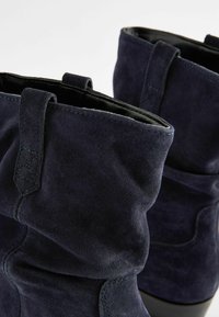 Tamsiai mėlynos suede kulkšninių botų su laisvąja forma, siuvinėjimo akcentais ir kilpuotais užtrauktukais, kad būtų lengva dėvėti.