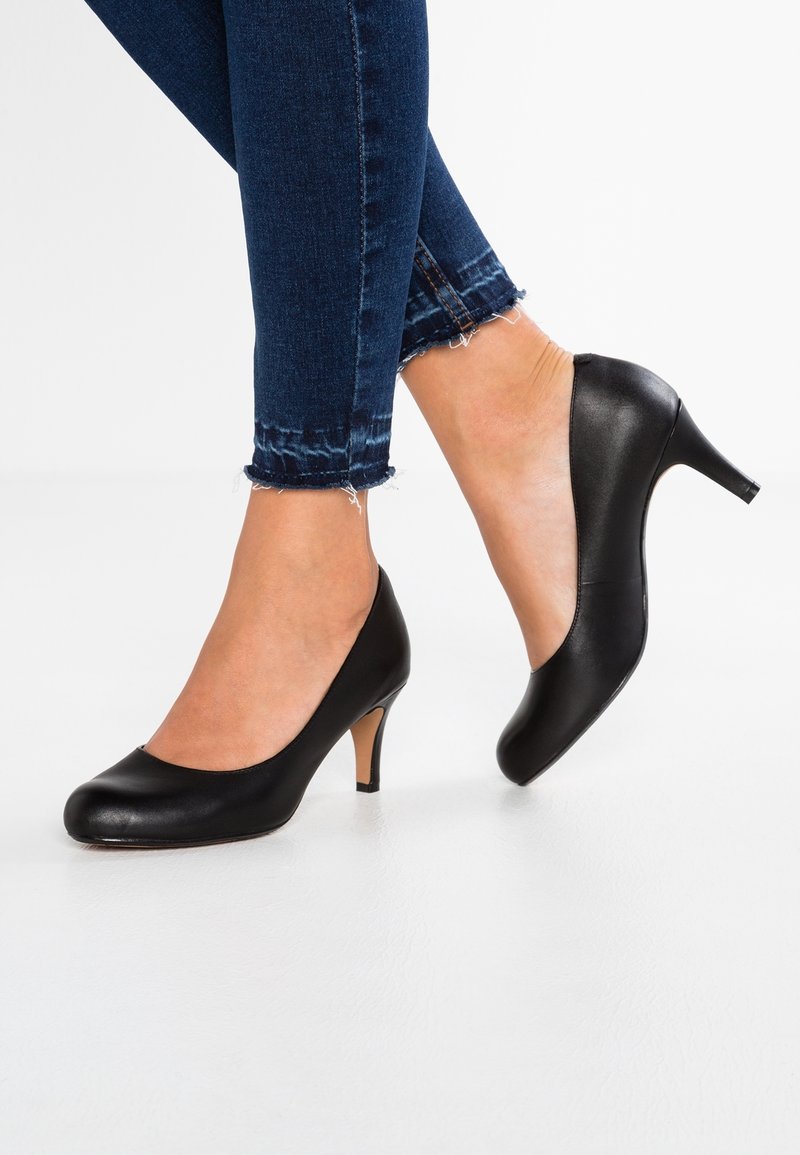 Pieds portant des escarpins en cuir noir à talon moyen, associés à un jean skinny bleu foncé à ourlet effiloché sur fond blanc.