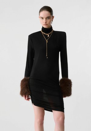 Femme portant une robe noire cintrée avec des poignets en fourrure marron, des manches longues et des colliers en chaînes dorées superposés, debout devant un fond clair uni.