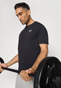 T-shirt noir en coton à manches courtes avec un logo blanc sur la poitrine, porté par une personne tenant une barre de poids. Design simple et ajusté.