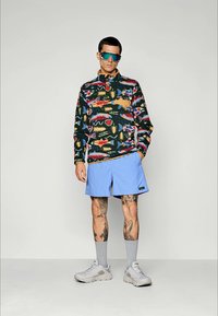 Multicolore fleece pullover met een patroon van vissen en abstracte vormen, gedragen met lichtblauwe shorts en grijze sokken, in combinatie met chunky witte sneakers.