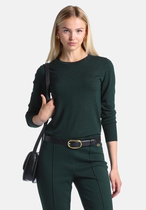 Femme blonde portant un pull vert foncé et un pantalon avec une ceinture noire et un sac à main, debout devant un fond clair.