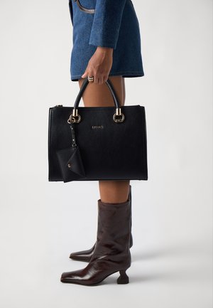 Borsa a tracolla in pelle nera con hardware dorato, forma strutturata e portacarte staccabile. Indossata con una giacca blu e stivali con tacco marroni.