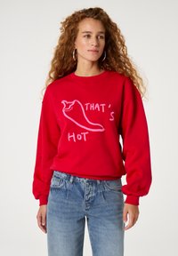 Roter Sweatshirt mit einer Chili-Pfeffer-Grafik und dem Text "DAS IST HEISS" in Pink. Das Design hat eine lässige Passform und gerippte Bündchen.