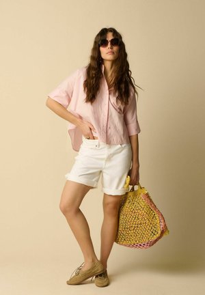Jeune femme aux longs cheveux ondulés portant une chemise rose oversize, un short blanc, des lunettes de soleil, des chaussures marron clair, tenant un sac fourre-tout tissé jaune et rose.