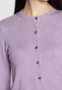 Cardigan en tricot lilas avec un col rond, une patte de boutonnage centrale avec des boutons brun foncé, et des détails côtelés au niveau du col.