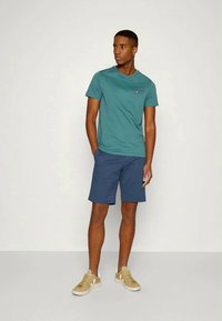 Jovem de pé com as mãos nos bolsos, vestindo uma t-shirt teal, calções azul-marinho e tênis bege contra um fundo claro e liso.