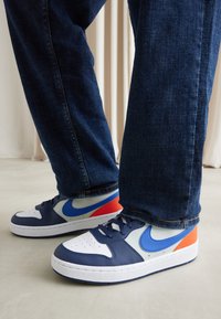 Navy and white sneakers with blue and orange accents, featuring leather upper, a perforated toe, and white rubber sole. Denim jeans visible.