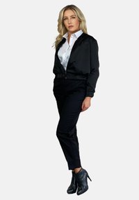 Czarna kurtka bomberka z zamkiem z przodu, haftowanym detalem, biała koszula pod spodem, czarne dopasowane spodnie i botki sięgające kostek. Gładki materiał i opływowy krój.