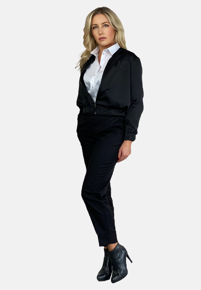 Czarna kurtka bomberka z zamkiem z przodu, haftowanym detalem, biała koszula pod spodem, czarne dopasowane spodnie i botki sięgające kostek. Gładki materiał i opływowy krój.