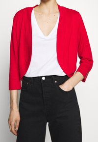Cárdigan corto rojo de material de punto ligero, con mangas tres cuartos y sutiles detalles de botones en los puños, usado sobre una camisa blanca.