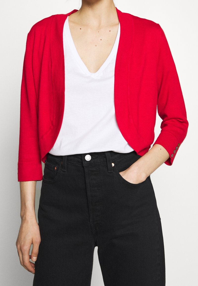 Cárdigan corto rojo de material de punto ligero, con mangas tres cuartos y sutiles detalles de botones en los puños, usado sobre una camisa blanca.