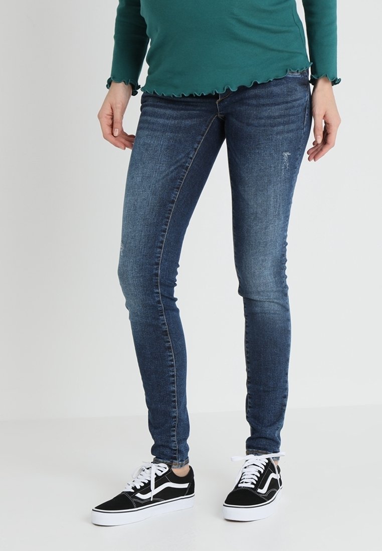 Jean skinny en denim bleu foncé, présentant un effet délavé, un style cinq poches et une coupe slim, associé à des baskets noires.