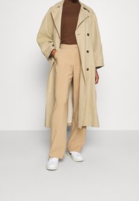 Trench-coat beige avec grands revers, boutons croisés, associé à un pantalon beige clair et des baskets blanches. Tissu lisse, coupe décontractée.