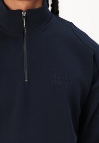 Marineblå hettejakke med høy krage, laget av strukturert stoff. Har en subtil brodert logo på brystet og metallglidelåsdetalj.