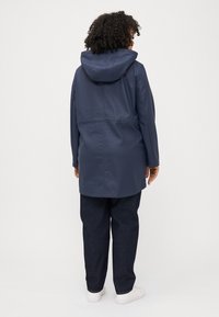 A woman with curly hair wears a navy raincoat with a hood, paired with dark jeans and white sneakers, viewed from the back.