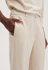 Beige op maat gemaakte broek van textuurstof, met zijzakken en een enkele knoopsluiting bij de taille. Subtiele stiksels zijn zichtbaar.