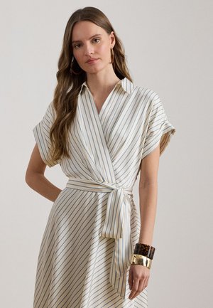 Hosszú hajú nő, fehér ruhában, amely vékony kék csíkokkal és megkötött derékövvel rendelkezik, nagy karika fülbevalókkal és vastag karkötőkkel kiegészítve.