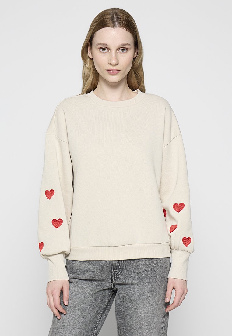 Frau in einem cremefarbenen Sweatshirt mit rotem Herzstickerei an den Ärmeln und grauen Jeans, die vor einem schlichten hellen Hintergrund steht.