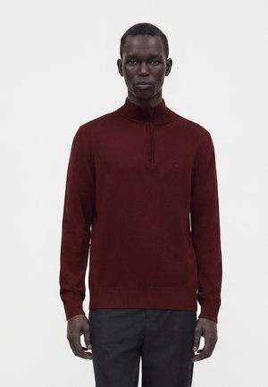 Homme portant un pull demi-zip rouge foncé à manches longues et un pantalon noir, debout devant un fond blanc uni.