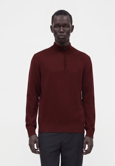 Homme portant un pull demi-zip rouge foncé à manches longues et un pantalon noir, debout devant un fond blanc uni.