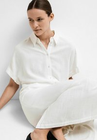 Chemise blanche à manches courtes, à coupe décontractée, fabriquée dans un tissu texturé. Associée à un pantalon léger assorti.