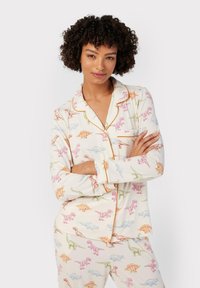 Pyjama van zachte stof met dinosaurusthema in crème. Voorzien van een kraag, knopen aan de voorkant en contrasterende oranje bies.
