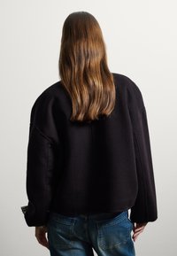 Pull noir oversize avec une texture douce et des épaules structurées, associé à un jean. Manches à carreaux visibles et coupe décontractée.