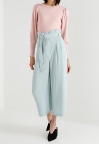 Haut rose clair à manches longues associé à un pantalon taille haute bleu clair à jambes larges, avec ceinture et texture en tissu. Chaussures noires à bout pointu.