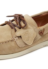 Beige Wildlederschuh mit braunen Schnürsenkeln, featuring ein lächelndes Charakteremblem und dekorativen Nähten. Flache Gummisohle mit strukturiertem Finish.